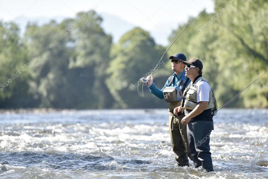 Guide demonstrating a fly cast beside a guest on a grassy riverbank.